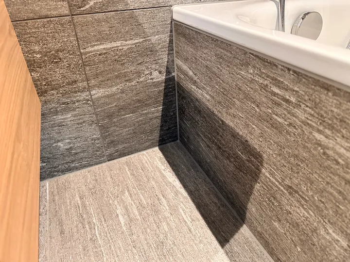 Badewanne mit Verkleidung aus grau gemaserten Platten in Steinoptik, Boden im gleichen Look, links eine helle Holzwand