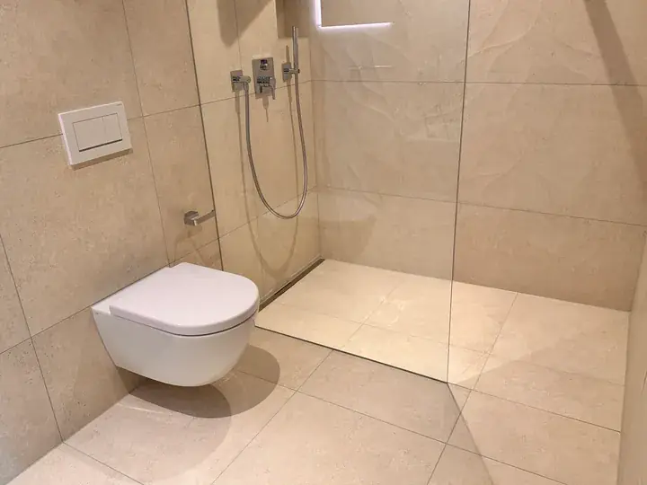 Badezimmer mit Dusche hinter einer Glasscheibe, Wand-WC und grossen beigen Platten an Wand und Boden