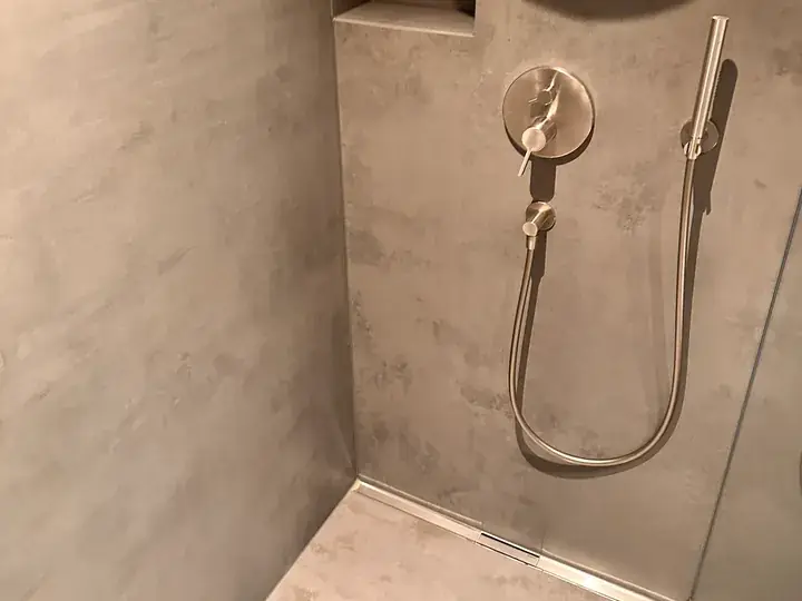 Dusche mit grau-beigen Wandflächen, Armatur mit Handbrause am Schlauch und eine Ablaufrinne am Boden