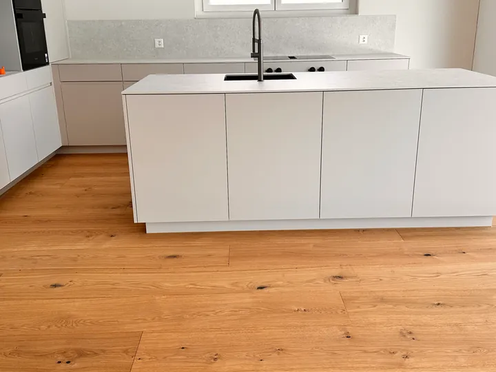 Helle Küche mit Insel, Spüle mit schwarzer Armatur und Holzboden, hinten eine helle Rückwand über der Arbeitsplatte