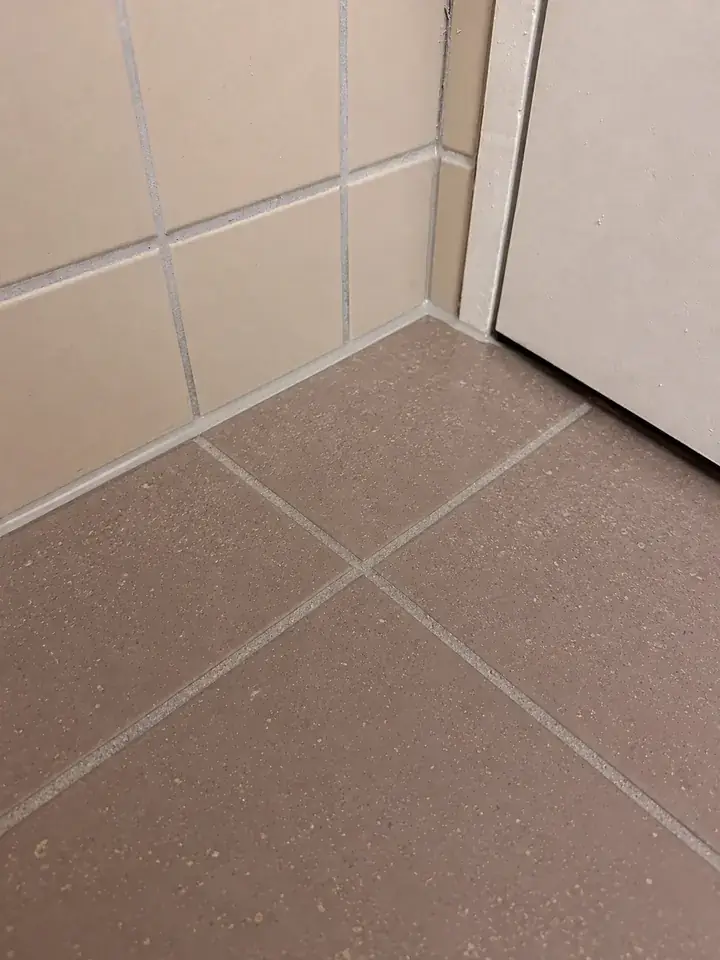 Dieselbe Ecke: Zwischen beigen Wandplatten und braunen Bodenplatten verläuft eine schmale helle Linie, rechts die Tür