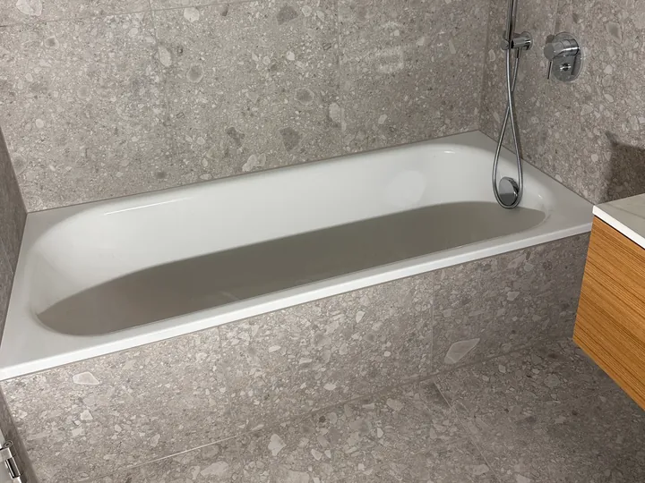Weisse Badewanne vor grauen Wandplatten mit Terrazzo-Muster, Handbrause und Armatur an der Wand, rechts ein Holzunterschrank