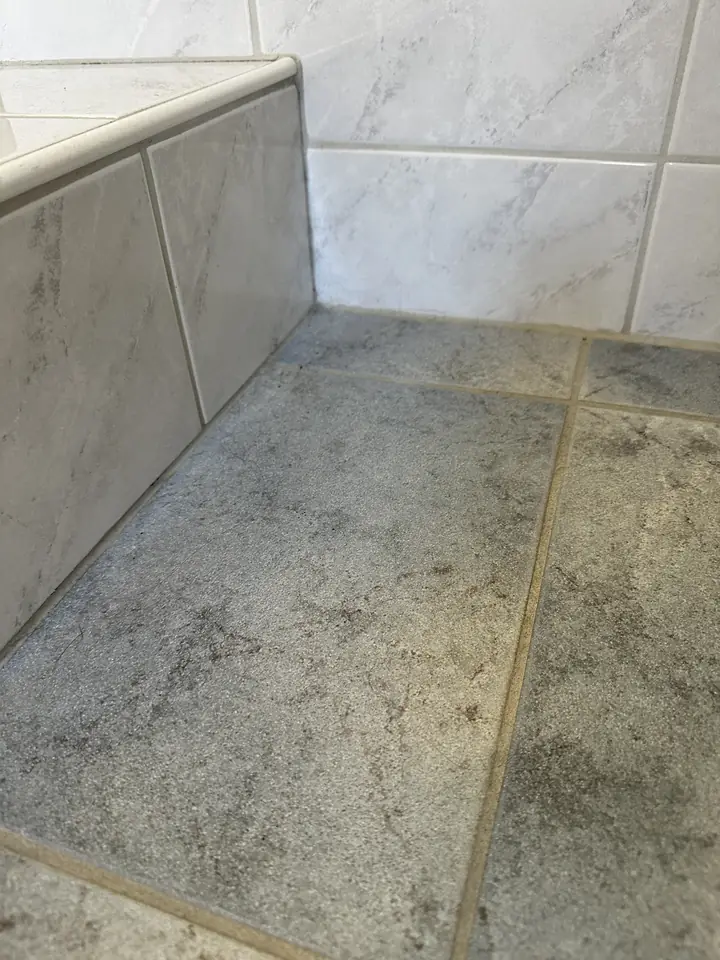 Grauer Fliesenboden vor weissen Wandplatten und gefliester Wannenverkleidung, entlang der Wand eine schmale graubraune Fuge