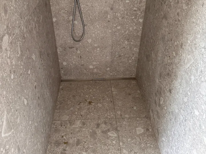 Schmale Dusche mit grau gesprenkelten Steinoptik-Platten an Wänden und Boden, Ablaufrinne an der Rückwand