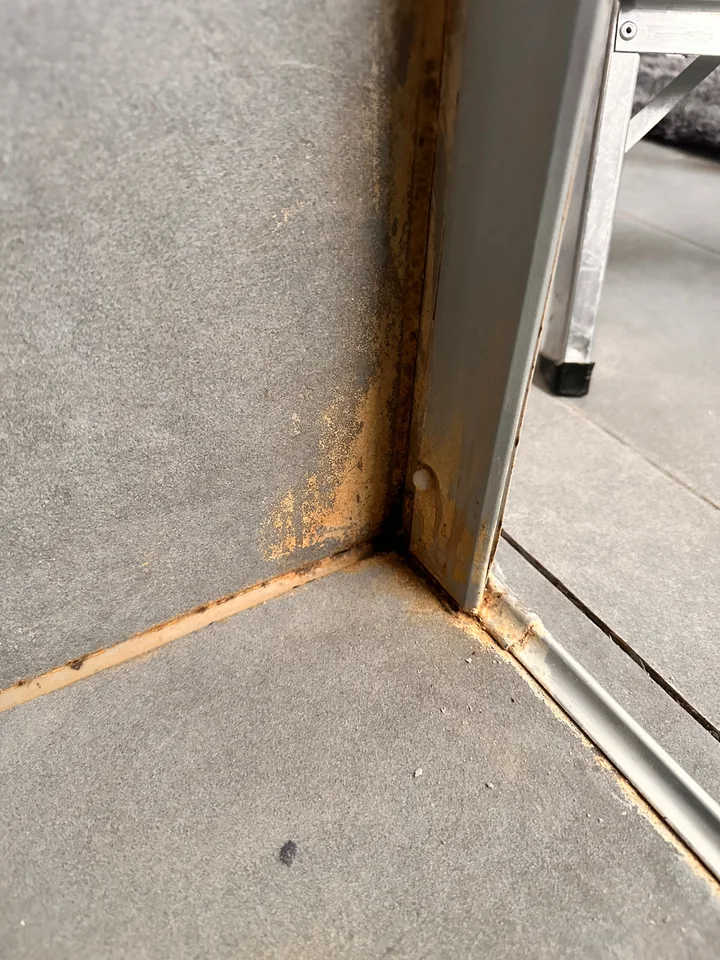 Ecke aus grauen Platten und Metallprofil, die Fuge am Boden und am Profil ist orangebraun verfärbt