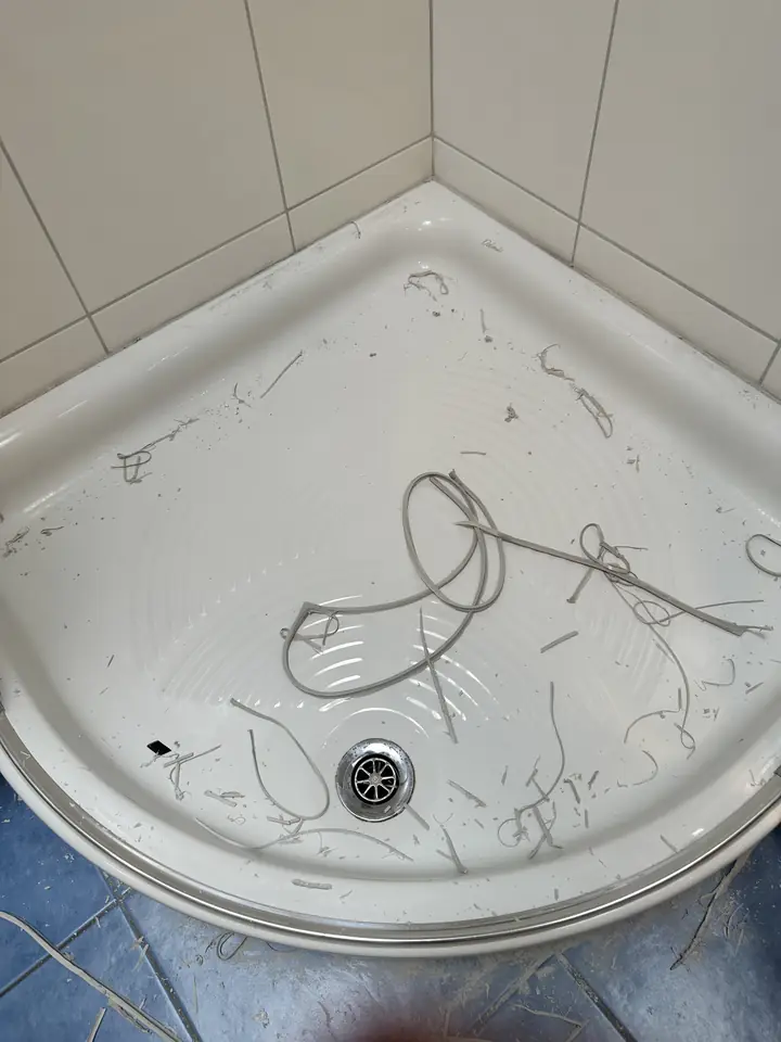 Weisse Eck-Duschwanne mit Ablauf vor hellen Wandplatten, in der Wanne liegen lose graue Silikonstreifen