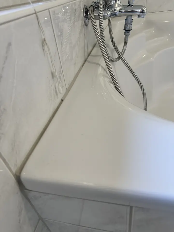 Weisse Eckbadewanne an marmorierten Wandplatten, die Fuge am Wannenrand ist dunkel, darüber Armatur mit Brauseschlauch