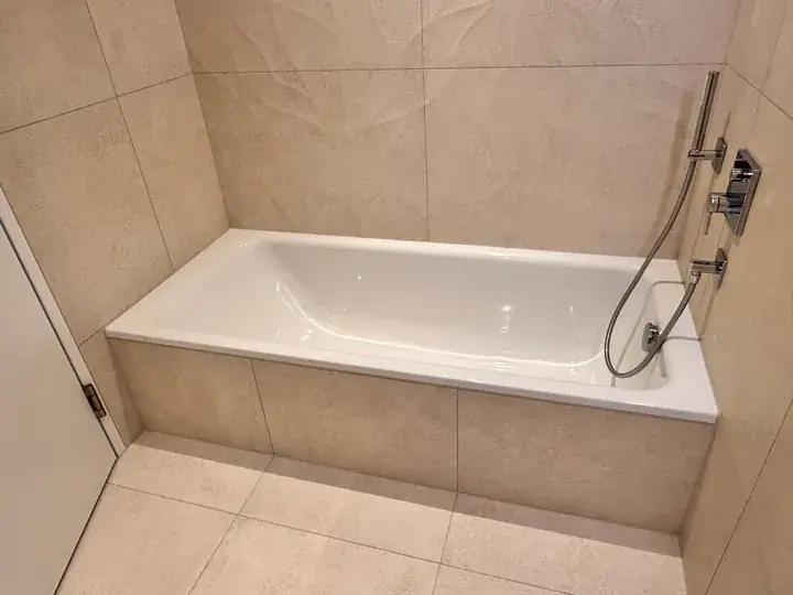 Eingebaute weisse Badewanne zwischen beigen Wandplatten, rechts Handbrause und Armatur, davor beige Bodenplatten