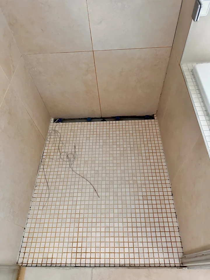 Duschboden aus kleinen hellen Mosaikfliesen zwischen beigen Wandplatten, hinten eine offene Leiste mit blauen Klammern