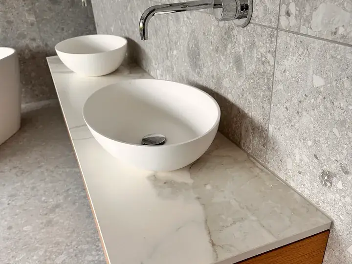 Zwei runde weisse Lavabos auf einer hellen Platte in Marmoroptik, dahinter Wandplatten in Steinoptik mit Wandarmatur