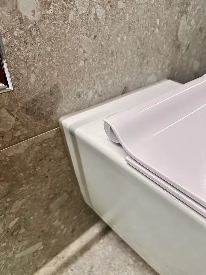 Weisses Wand-WC mit Deckel vor Wandplatten in Steinoptik, links am Bildrand ein Stück Chrom