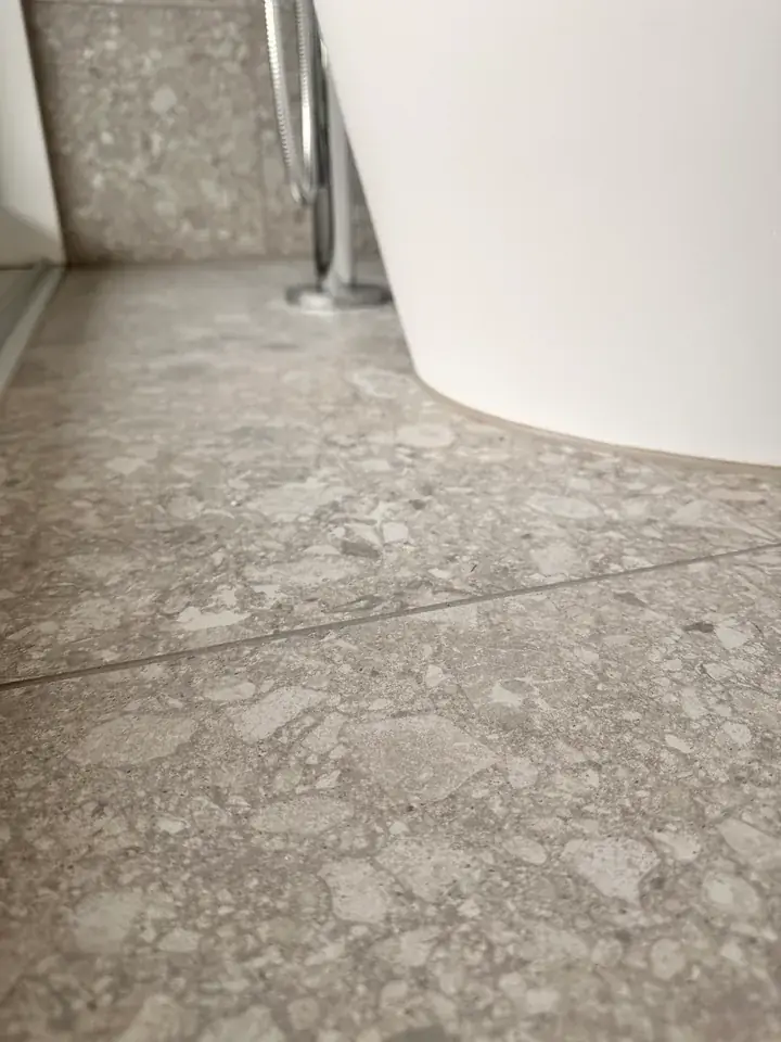 Weisse, runde Badewanne auf einem Boden aus Platten in grau-beiger Steinoptik, daneben eine verchromte Armatur