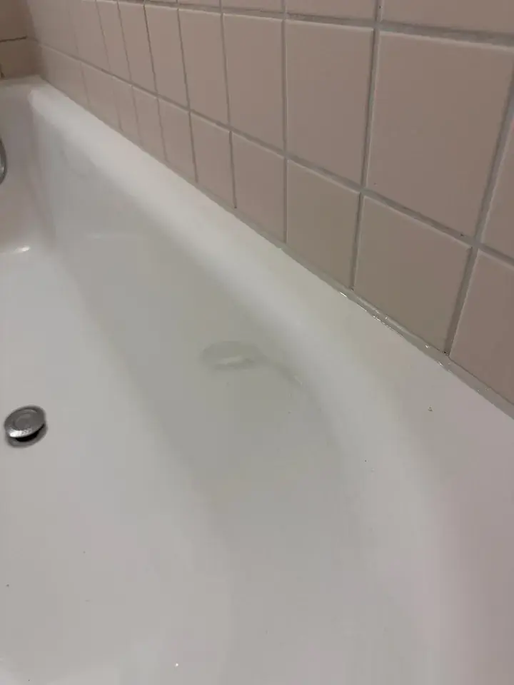 Weisse Badewanne vor rosabeigen Wandplatten, an der Kante zur Wand verläuft eine schmale helle Linie, links der Ablauf