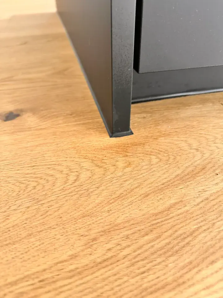 Holzboden mit Maserung an der Ecke eines schwarzen Möbelkorpus, unten am Korpus eine schmale dunkle Kante
