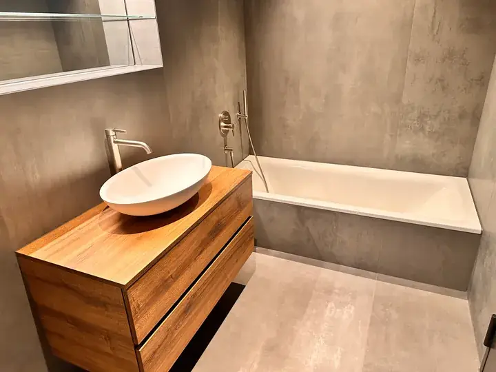 Badezimmer mit weisser Badewanne, ovalem Aufsatzbecken auf Holzunterschrank und grauen Grossformatplatten an Wand und Boden