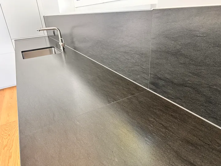 Dunkle Küchenarbeitsplatte mit Spüle und Armatur vor einer dunklen Rückwand; zwischen beiden verläuft eine helle Linie
