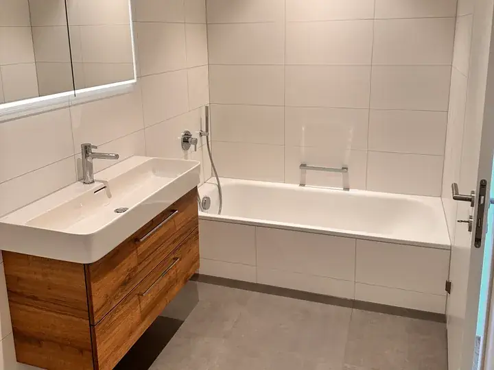 Helles Badezimmer mit Badewanne, Waschtisch mit Holzunterschrank, grossen weissen Wandplatten und grauem Boden