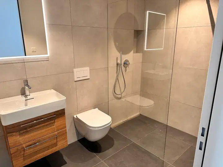 Badezimmer mit Glaswand an der Dusche, wandhängendem WC, Waschtisch mit Holzunterschrank und beigen Wandplatten