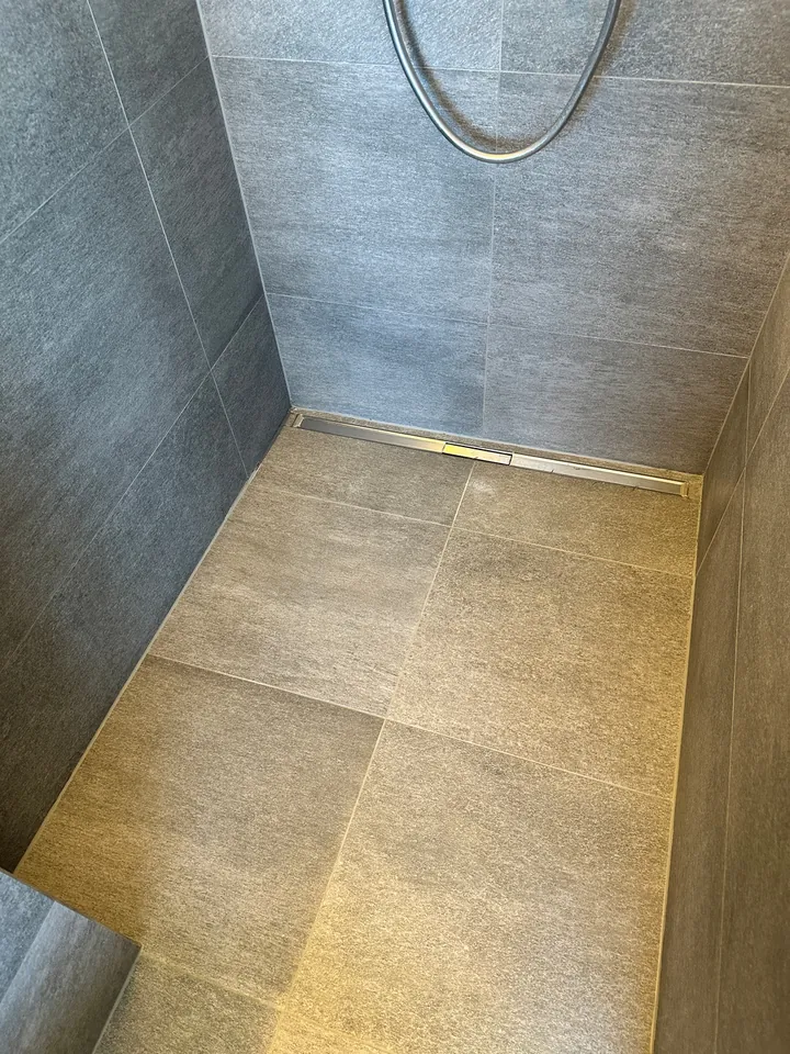 Dusche mit grauen Wandplatten, beigem Boden und Metall-Ablaufrinne, am Rand zwischen Boden und Wand ein heller Streifen