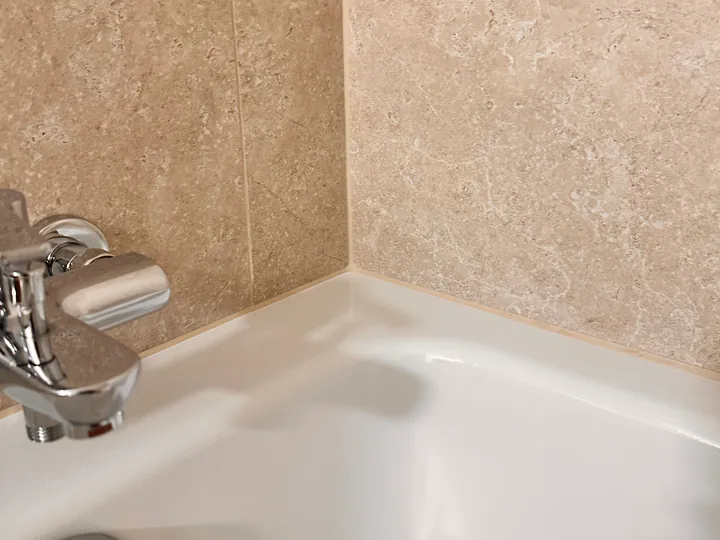 Ecke einer weissen Badewanne neben der Armatur, beige Wandplatten mit heller Fuge am Wannenrand