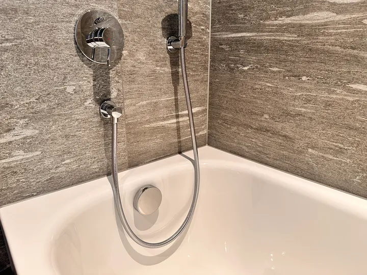 Weisse Badewanne mit Einhebelmischer und Handbrause vor grauen Wandplatten in Steinoptik