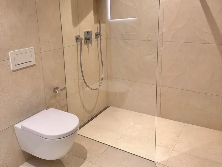Bodengleiche Dusche mit Glaswand, beigen Grossformatplatten und Handbrause, links ein wandhängendes WC mit Spülplatte