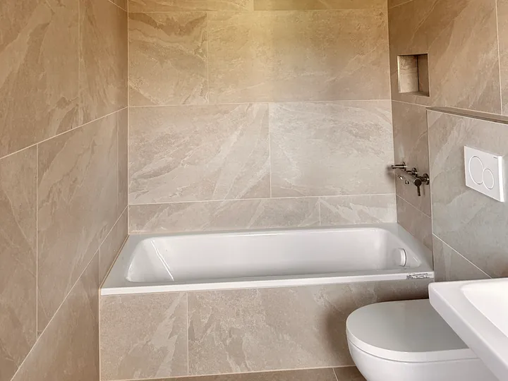 Eingebaute weisse Badewanne vor grossformatigen Wandplatten in Steinoptik, rechts ein Wand-WC und der Rand eines Waschtischs