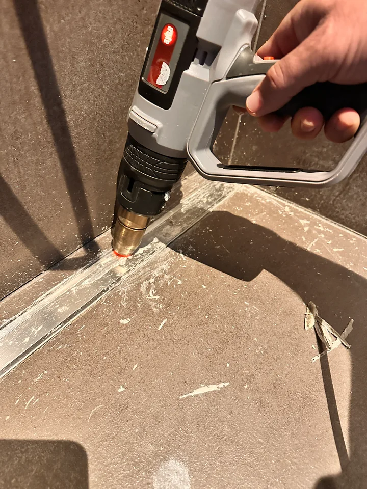 Hand hält ein graues Gerät mit Düse an ein Metallprofil im Duschboden, auf den Platten liegen Silikonreste