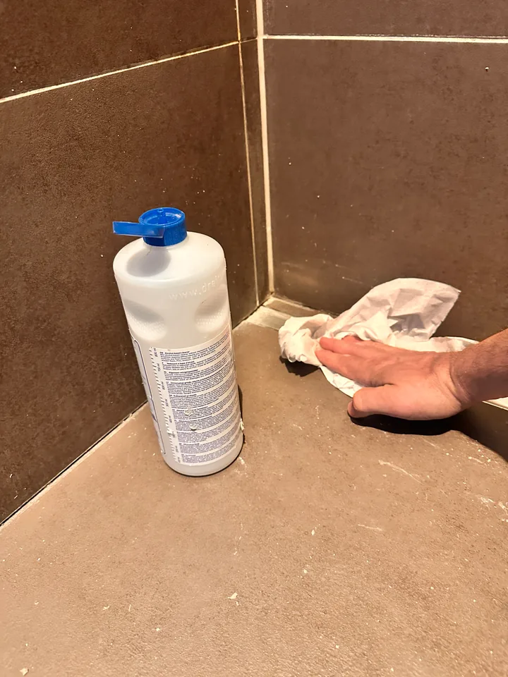 Weisse Flasche Reinigungsmittel steht in einer Ecke der Dusche, eine Hand wischt mit einem Papiertuch am Boden