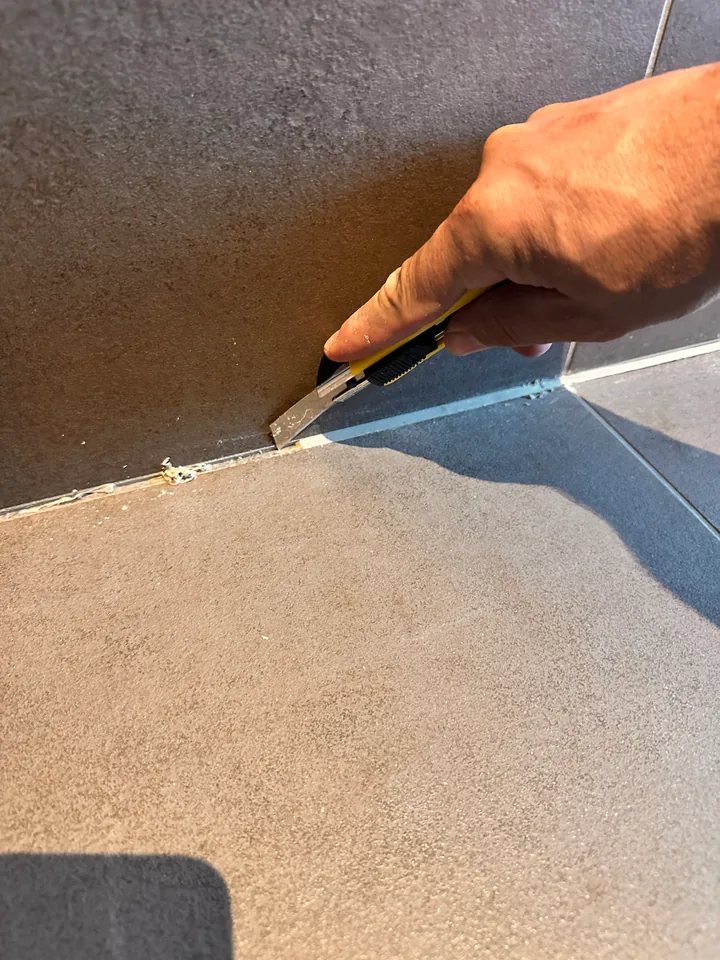 Hand mit gelbem Cuttermesser an der Kante zwischen dunkler Wandplatte und Boden, am Rand Reste von altem Silikon