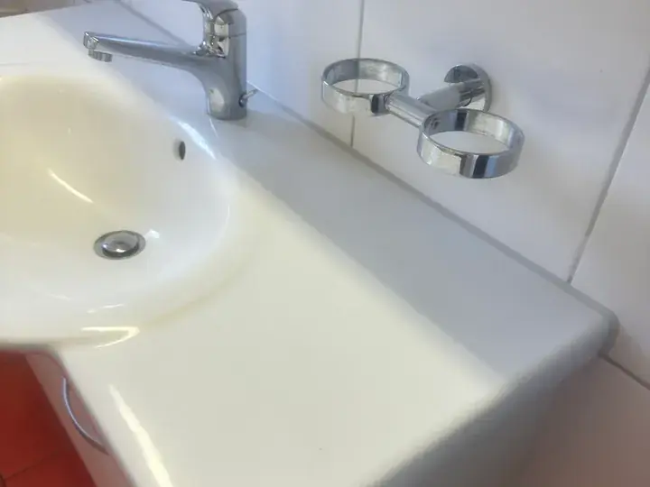 Weisser Waschtisch mit Armatur und Becherhalter vor weissen Wandplatten, unten links ein roter Schrank