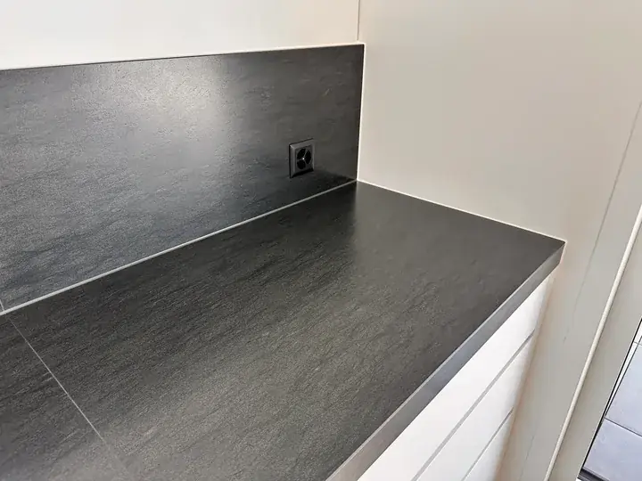 Dunkle Arbeitsplatte mit dunkler Plattenrückwand und Steckdose vor weisser Wand, rechts weisse Schrankfronten