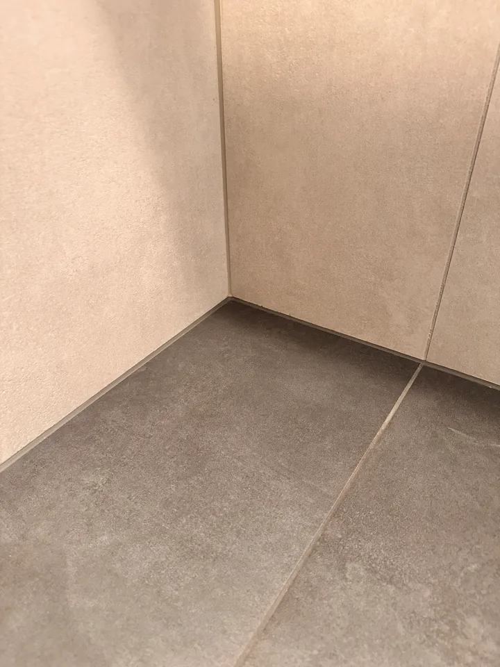 Graue Bodenplatten an beigen Wandplatten in einer Ecke, entlang der Kante zur Wand verläuft eine schmale Fuge
