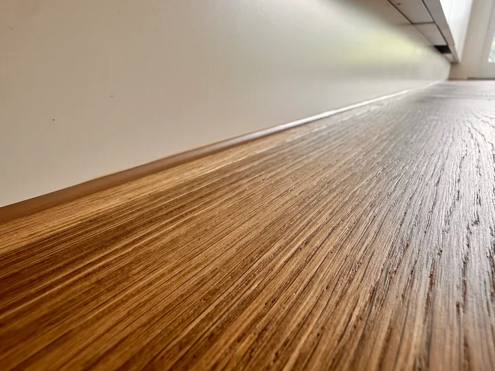 Holzboden mit Längsmaserung an einer weissen Wand, am Übergang verläuft ein schmaler hellbrauner Streifen