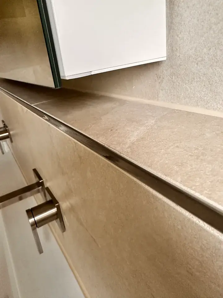 Lange Ablage mit beigem Plattenbelag und Metallkante, darüber ein Spiegelschrank, links Armaturgriffe aus Metall