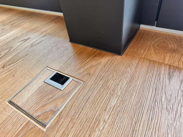 Holzboden mit eingelassener Bodensteckdose neben einem dunklen, eckigen Pfosten an der Wand