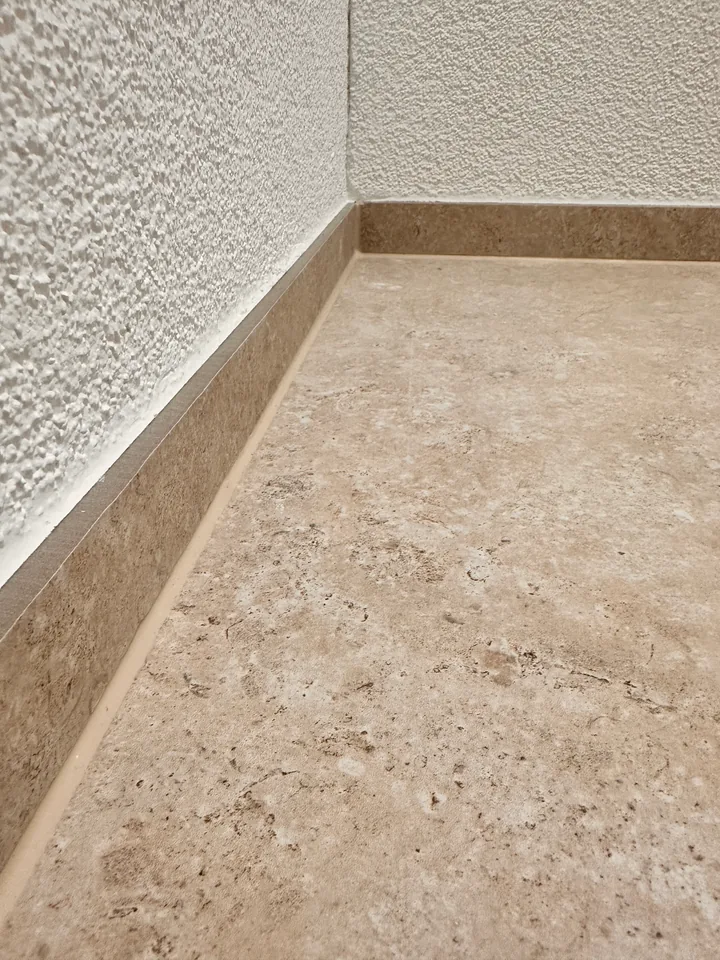 Beiger Plattenboden mit Sockelleiste im selben Beigeton vor weisser, rau strukturierter Wand