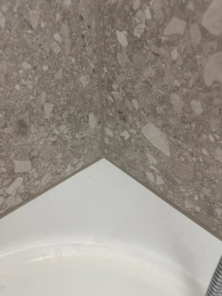 Innenecke aus grauen Wandplatten mit Steinmuster über einer weissen Wanne, unten rechts ein Metallschlauch
