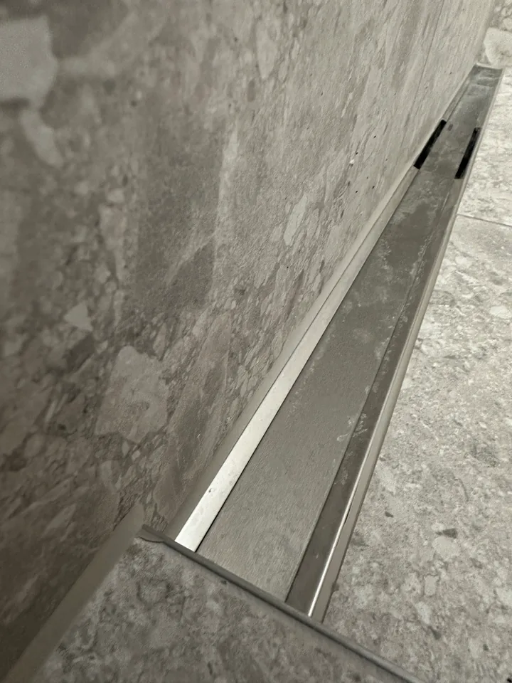 Duschrinne aus Edelstahl entlang einer Wand aus grauen Platten mit Steinmuster, rechts graue Bodenplatten