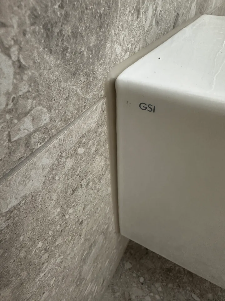Weisser Sanitärkörper seitlich an einer grauen Wand mit Steinmuster, an der Kante entlang eine schmale helle Fuge