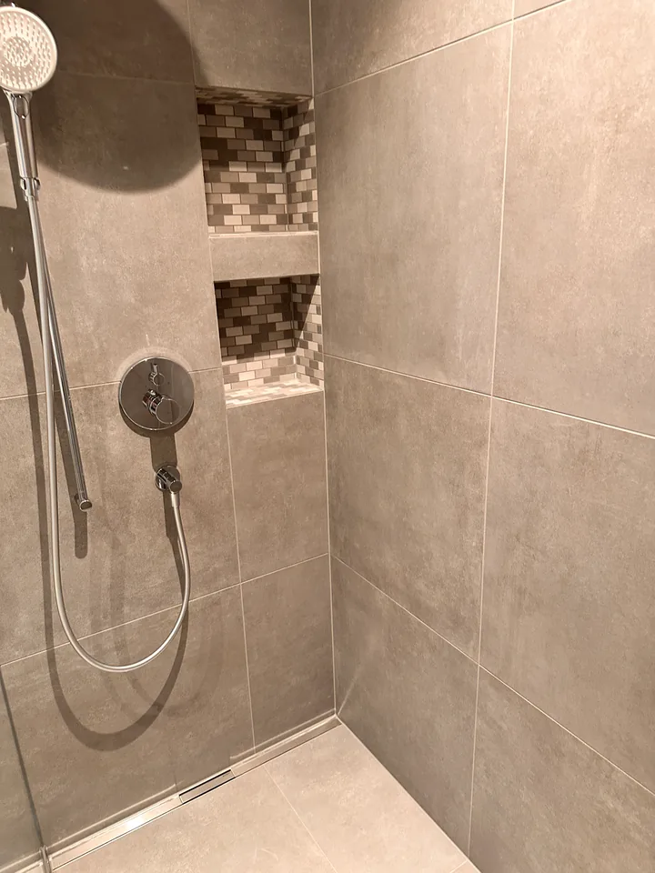 Dusche mit grossen beigegrauen Wandplatten, Handbrause mit Schlauch an der Stange, Mosaiknische und Ablaufrinne am Boden