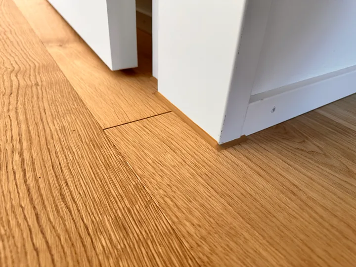 Eichenparkett an einem weissen Rahmen mit Leiste, in der Ecke ein schmaler brauner Streifen am Parkettrand