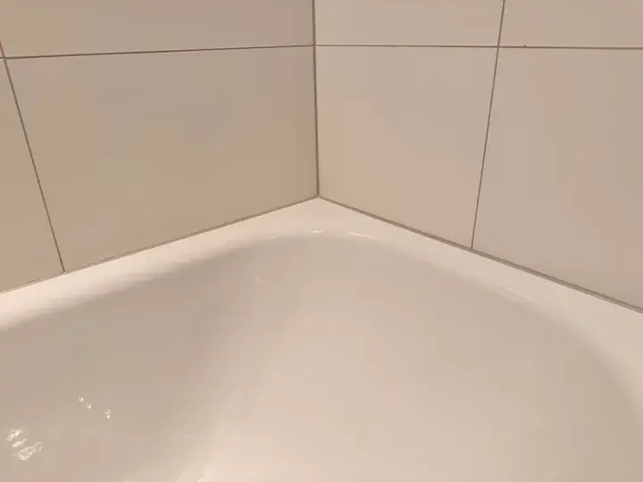 Ecke einer weissen Badewanne an hellen, grossformatigen Wandplatten, am Wannenrand verläuft eine dünne graue Fuge