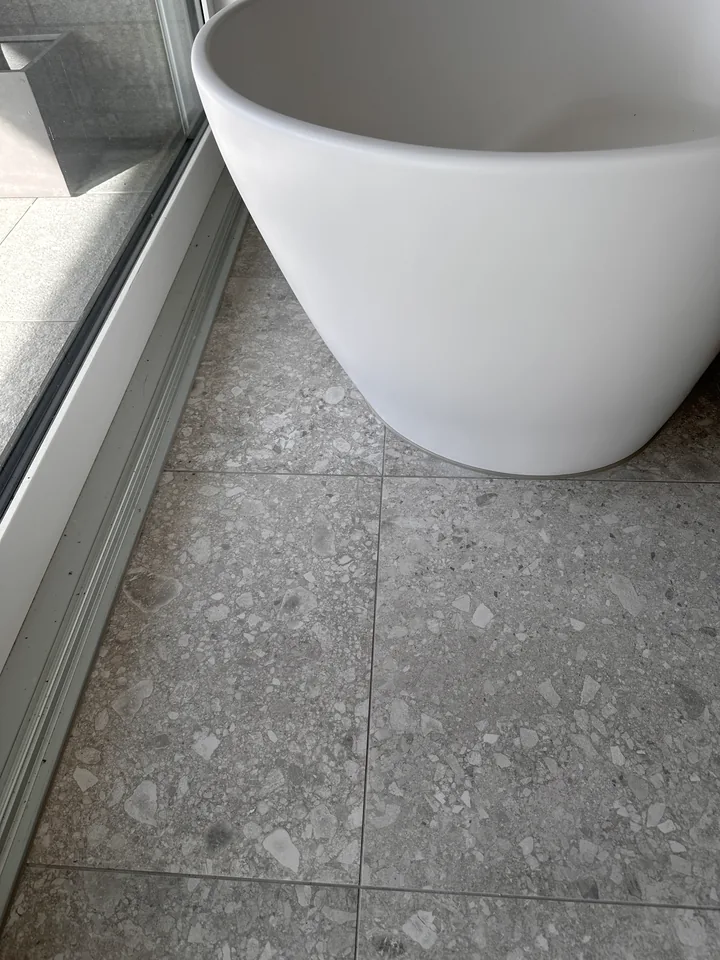 Freistehende weisse Badewanne auf grauem Fliesenboden, links ein Glaselement mit hellem Rahmen