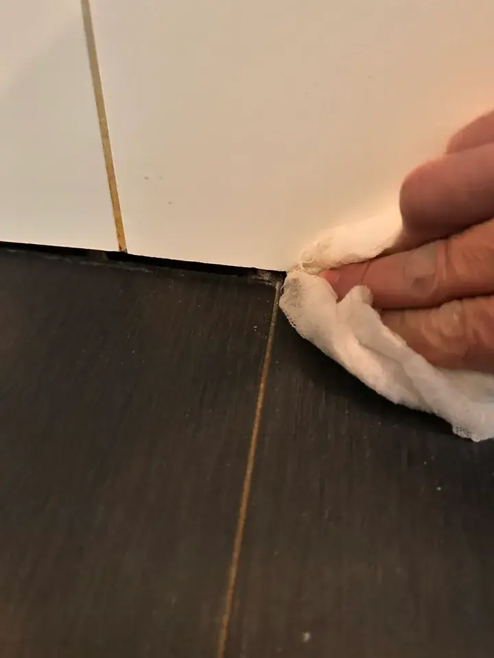 Hand mit weissem Tuch am Übergang von hellen Wandplatten zum dunklen Boden, daneben eine Bodenfuge