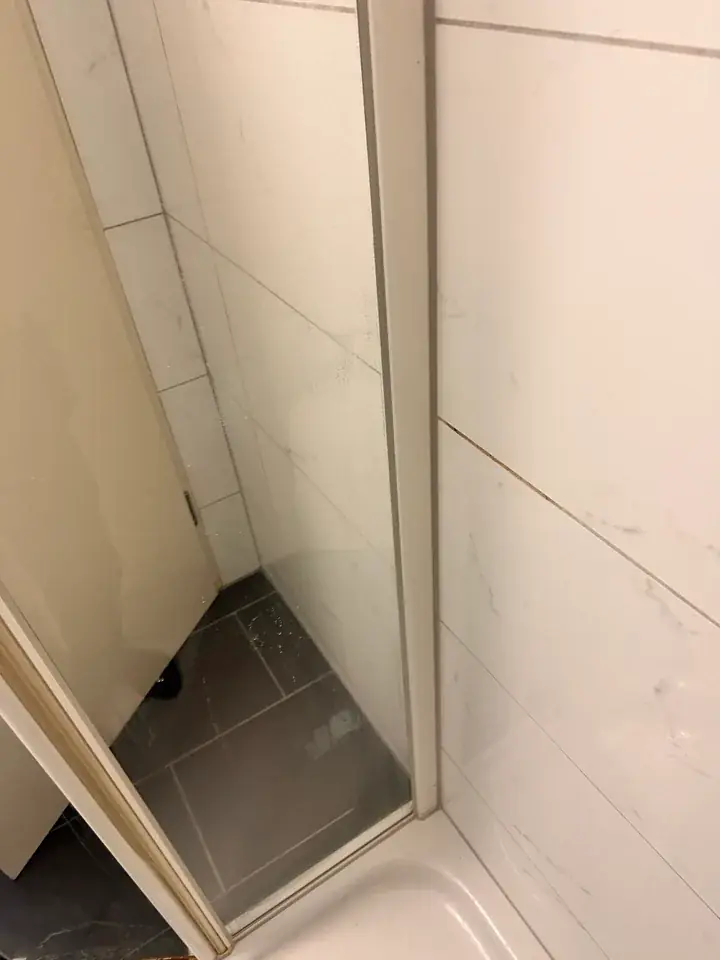 Senkrechtes weisses Profil einer Duschkabine mit Glas neben weissen Wandplatten, unten der Rand einer weissen Duschtasse