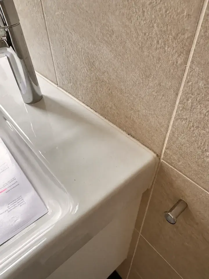 Weisses Lavabo mit Chromarmatur vor beigen Reliefplatten, am Übergang zur Wand ein heller Streifen mit dunklen Punkten