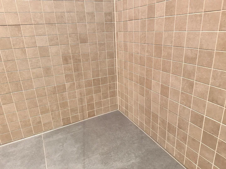 Duschecke mit beigen Mosaikfliesen an zwei Wänden, am Übergang zum grauen Boden verläuft eine helle Fuge