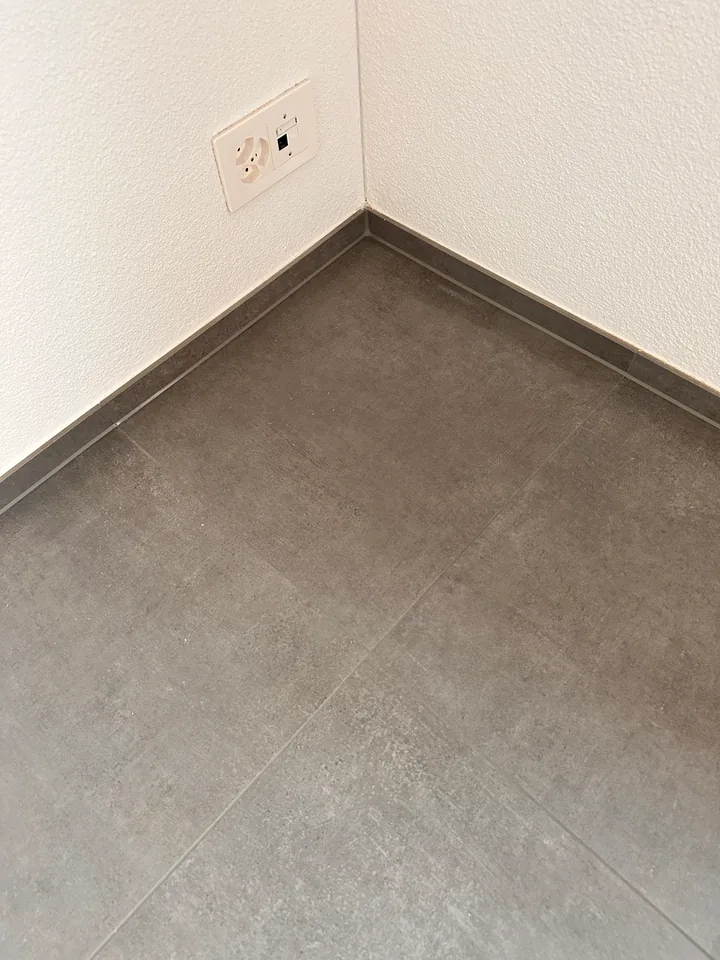 Raumecke mit weisser Wand und Steckdose, grauen Bodenplatten und grauer Sockelleiste, dazwischen eine schmale Fuge