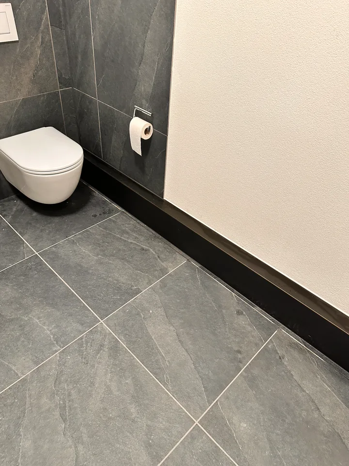 Weisses Wand-WC vor dunkelgrauen Wandplatten, dazu Plattenboden und eine schwarze Sockelleiste an einer weissen Wand